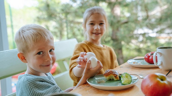 Geschwister halten Eier und genießen das Frühstück im Bauernhaus.
