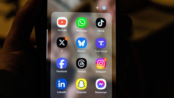 Smartphone-Bildschirm mit dem Logo von Bluesky neben dem Logo von „X“ und dem Logo von Donald Trumps Truth Social-App. In der oberen Reihe von links nach rechts ist das Logo von YouTube neben dem von WhatsApp und TikTok zu sehen. In der unteren Reihe von links nach rechts sind die Logos für Facebook, Threads, Instagram, LinkedIn, Snapchat und Messenger zu sehen.