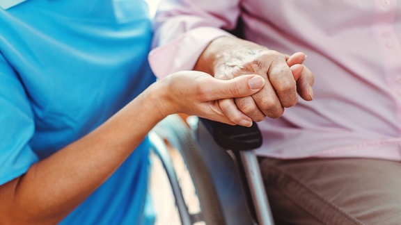 Eine Pflegerin hält die Hand eines Menschen im Rollstuhl