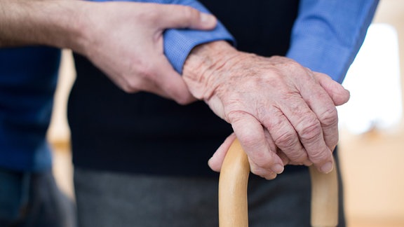 Nahaufnahme wie sich eine ältere Person auf einen Gehstock stützt und dabei von einer jungen Person am Arm gehalten wird.