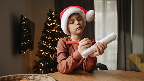 Ein Junge mit einer Weihnachtsmütze schreibt auf ein Papier.