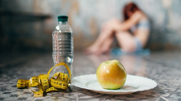 Frau sitzt im Hintergrund am Boden. Im Vordergrund ein Apfel, eine Flasche Wasser und ein Maßband.
