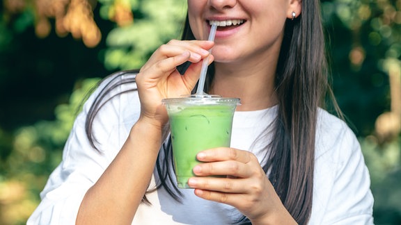 Eine junge Frau trinkt eine grünes Getränk aus einem umweltschädlichen Plastikbecher mittels Wegwerfstrohhalm. 