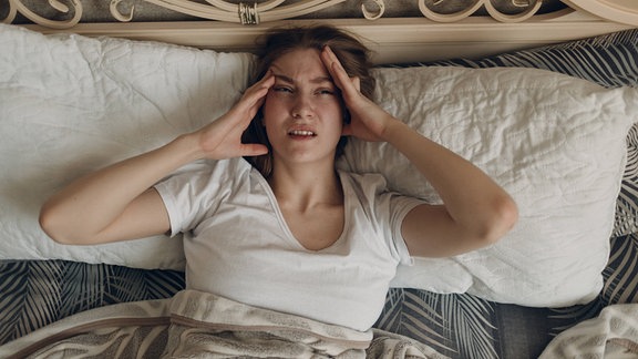 Eine Frau liegt im Bett und hält sich den Kopf.
