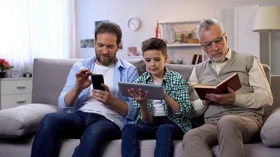 Ein älterer Mann liest ein Buch, ein mittelalter Mann schaut auf ein Handy und ein Junge mit einem Tablet