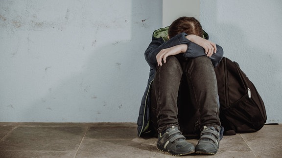 Ein Kind mit Schulrucksack sitzt am Boden und legt den Kopf auf die Unterarme.
