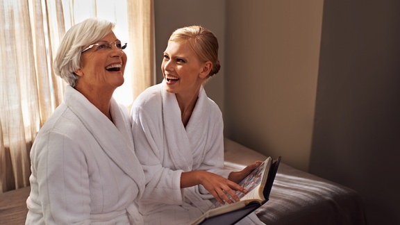 Zwei Frauen in Bademäntlen sitzen nebeneinander und lachen. 