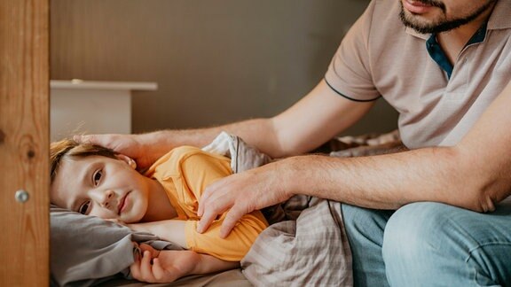 Ein Kind liegt im Bett während der Vater ihm den Kopf streichelt.