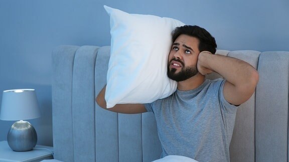 Unglücklicher junger Mann hält sich im Bett die Ohren zu.