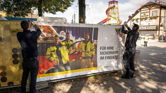 Mitarbeiter der Feuerwehr bringen ein Banner mit der Aufschrift Für Ihre Sicherheit im Einsatz an einem Zaun an