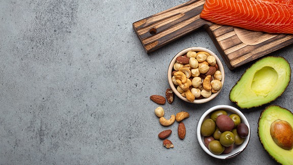 Nahrungsquellen für gesunde ungesättigte Fette und Omega 3: frisches rohes Lachsfilet, Avocado, Oliven, Nüsse auf Schneidebrett, rustikaler Steinhintergrund von oben.
