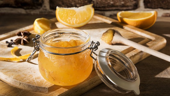 Offenes Glas Orangenmarmelade auf einem Brett mit Orangenstücken, Gewürzen und Holzlöffel