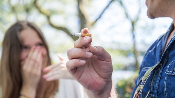 Ein Mann raucht in einem Park eine Zigarette und stört damit die Frau, die neben ihm sitzt (gestellte Szene).