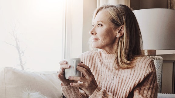 Eine Frau mittleren Alters hält eine Tasse und blickt aus dem Fenster hinaus.