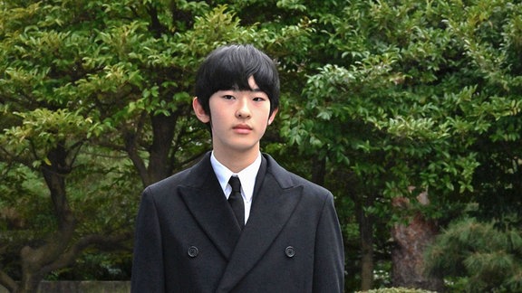 Prinz Hisahito von Japan in einem eleganten schwarzen Mantel in einem Park laufend.
