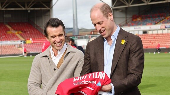 Rob McElhenney, Vorsitzender des Wrexham AFC, und Prinz William, Prinz von Wales, mit seinem personalisierten Wrexham AFC-Trikot auf dem Spielfeld des Racecourse Ground.