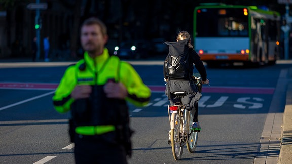 Momentaufnahme aus dem Straßenverkehr: Ein Polizist im Bildvordergrund, dahinter ein*e Radfahrer*in, dahinter ein Bus