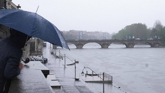 Eine Person mit Regenschirm schaut auf einen Fluss, der Hochwasser führt.