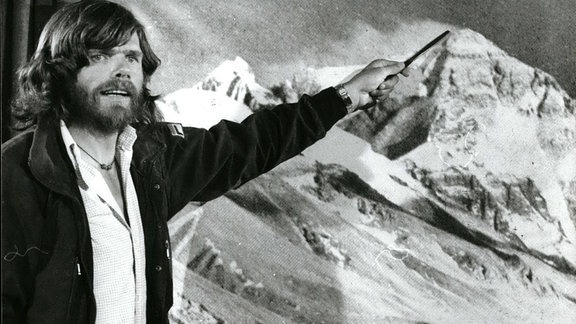 Reinhold Messner zeigt auf eine Bergspitze