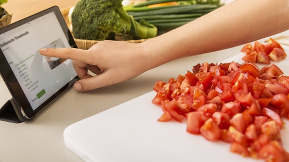 Eine junge Frau schneidet Tomaten auf dem Küchentisch und liest ein Rezept auf einem Tablet.