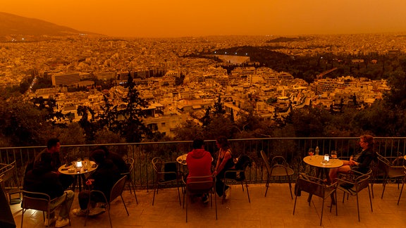 Gäste sitzen auf der Terrasse eines Restaurants mit Blick vom Filopappou-Hügel, über allem liegt ein orangener Schleier.