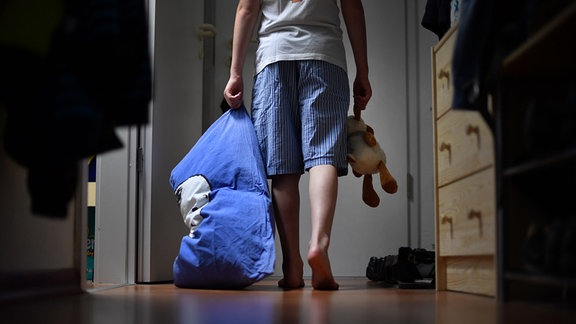Ein Junge im Schlafanzug geht mit einem Kopfkissen und einem Kuscheltier in der Hand durch einen Flur zur Wohnungstür