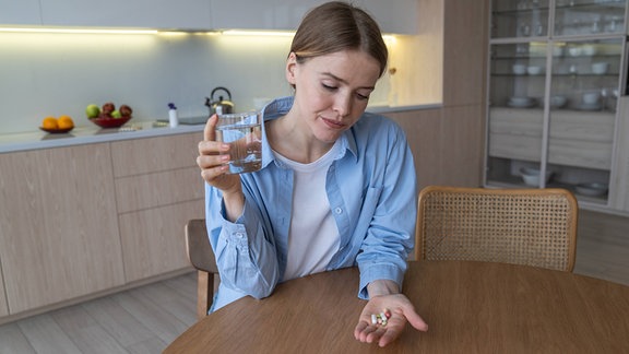 Unzufriedene, grimassierende Frau schluckt Tabletten gegen Kopfschmerzen und Übelkeit und fühlt sich am Küchentisch unwohl