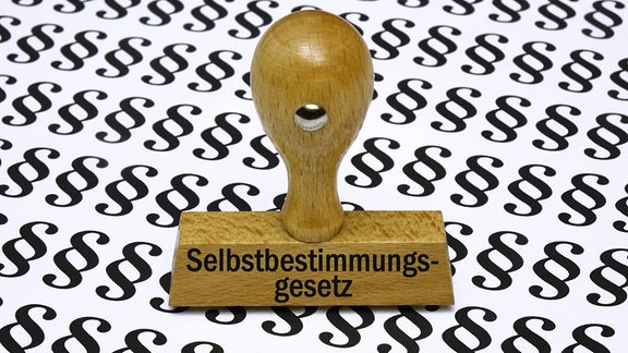 Symolbild: Hölzerner Stempel mit Aufschrift "Selbstbestimmungsgesetz" auf Paragrafenpapier