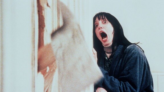 Szene aus dem Horrorfilm "The Shining": eine Frau lehnt an der Wand und schreit, weil eine Axt die Wand durchschlägt