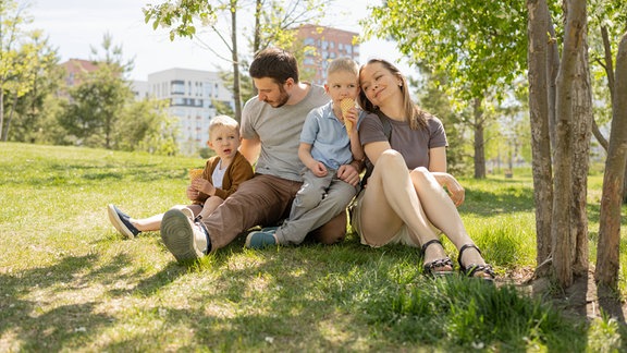 Eine vierköpfige Familie, bestehend aus Mutter, Vater und zwei kleinen Söhnen, entspannt im Schatten eines blühenden Baums in einem üppigen Park.