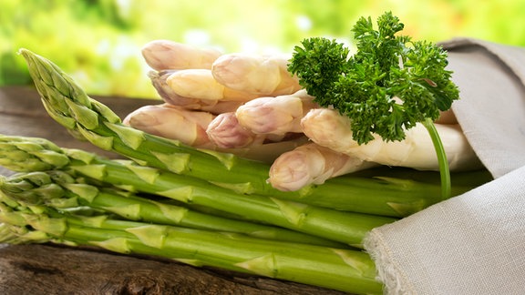 Zwei Bund, gründer und weißer Spargel, liegen mit Petersilie auf einem Tisch.