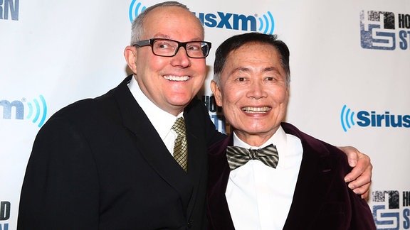 George Takei (rechts) und Ehemann Brad Takei umarmen sich.