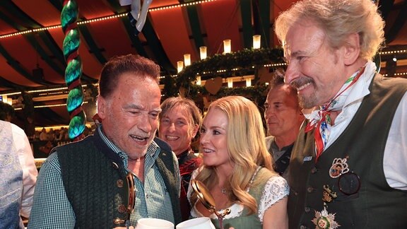 Arnold Schwarzenegger (l-r), Schauspieler, Schwarzeneggers Partnerin Heather Milligan und Thomas Gottschalk, Entertainer, stehen im Marstall-Festzelt auf der Bühne. Die Wiesn findet vom 21. September bis 6. Oktober 2024 statt.
