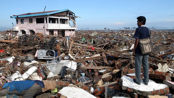 Ein Mann steht zwischen den Trümmern seines Hauses und Heimatortes in Banda Aceh.