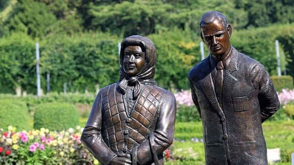 Zwei festlich gekleidete Männer enthüllen eine Statue zum Gedenken an Königin Elisabeth II.