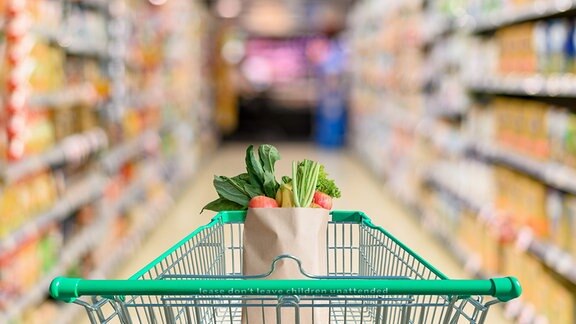 Inszeniertes Foto eines Einkaufswagens, in dem eine Papiertüte Gemüse steht und der im Gang eines Supermarktes steht.