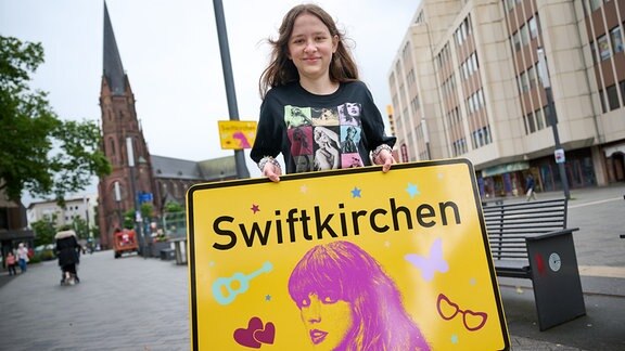 Die Schülerin Aleshanee Westhoff zeigt im Anschluss an offizielle Einweihung auf dem Heinrich-König-Platz in Gelsenkirchen das Ortsschild "Swiftkirchen" zu Ehren der Musikerin Taylor Swift.