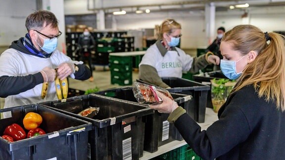 In einer Halle packen ehrenamtliche Helfer der Berliner Tafel mit Atemschutzmasken Lebensmittel in Tüten