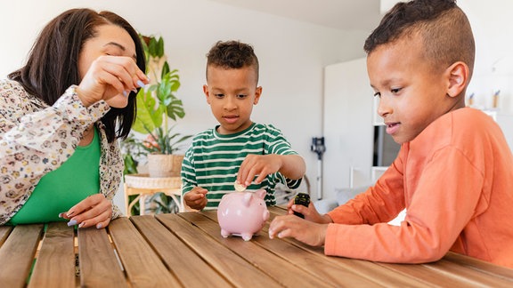 Eine Mutter und zwei Jungen werfen zu Hause Münzen ins Sparschwein.