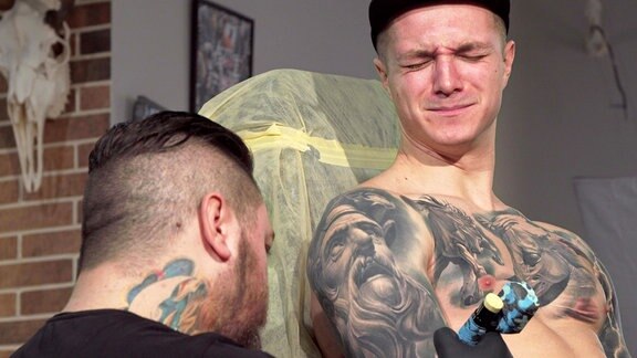 Junger Mann lässt sich schmerzhaft ein Tattoo auf den Arm machen. Rückansicht eines professionellen Tätowierers bei der Arbeit.