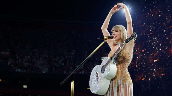 Taylor Swift macht auf einer Bühne ein Herz mit ihren Händen.