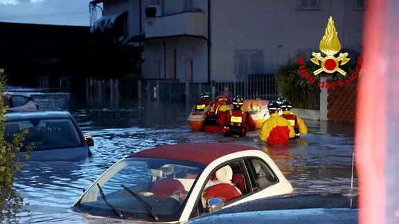 Feuerwehrleute schieben ein Schlauchboot durch eine überschwemmte Straße, in der zahlreiche Autos unter Wasser stehen.
