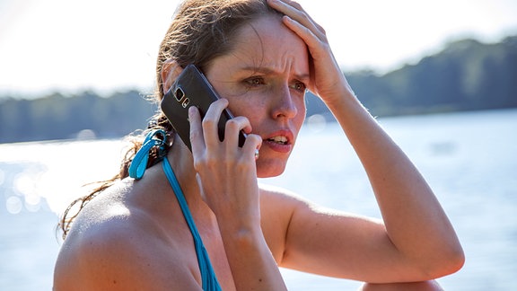 Eine Frau an einem Badesee sitzend,  telefoniert mit angestrengtem Blick, mit ihrem Smartphone.