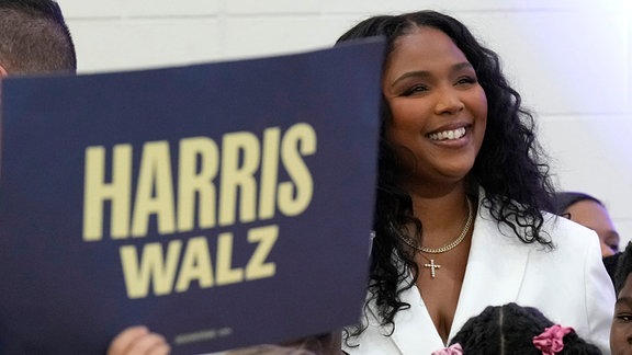Sängerin Lizzo steht lachend hinter einem Plakat mit der Aufschrift "Harris, Walz".