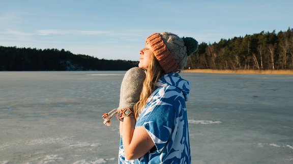 Eine junge Frau steht an einem See und richtet sich mit geschlossenen Augen und vor der Brust gefalteten Händen in Richtung Sonne.