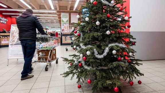 Mann mit Einkaufswagen neben Weihnachtsbaum in Supermarkt.