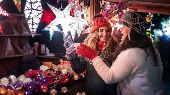 Zwei freudige junge Frauen betrachten weihnachtliche Dekoartikel an einem Stand auf dem Weihnachtsmarkt.