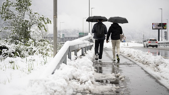 Zwei Personen gehen auf einem teilweise vom Schnee geräumten Bürgersteig auf einer Brücke entlang.