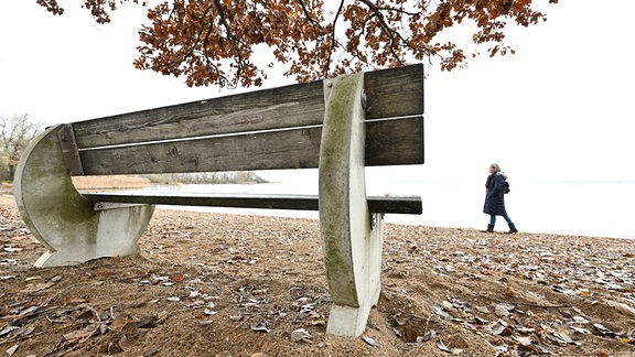 Leere Sitzbank, dahinter Person am See
