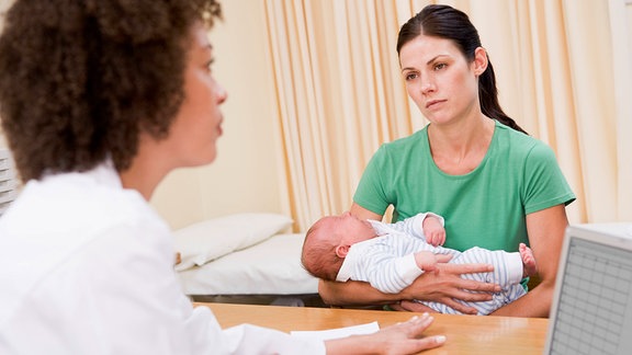 Einer Ärztin am Laptop sitzt eine Frau gegenüber, die ein Baby hält.